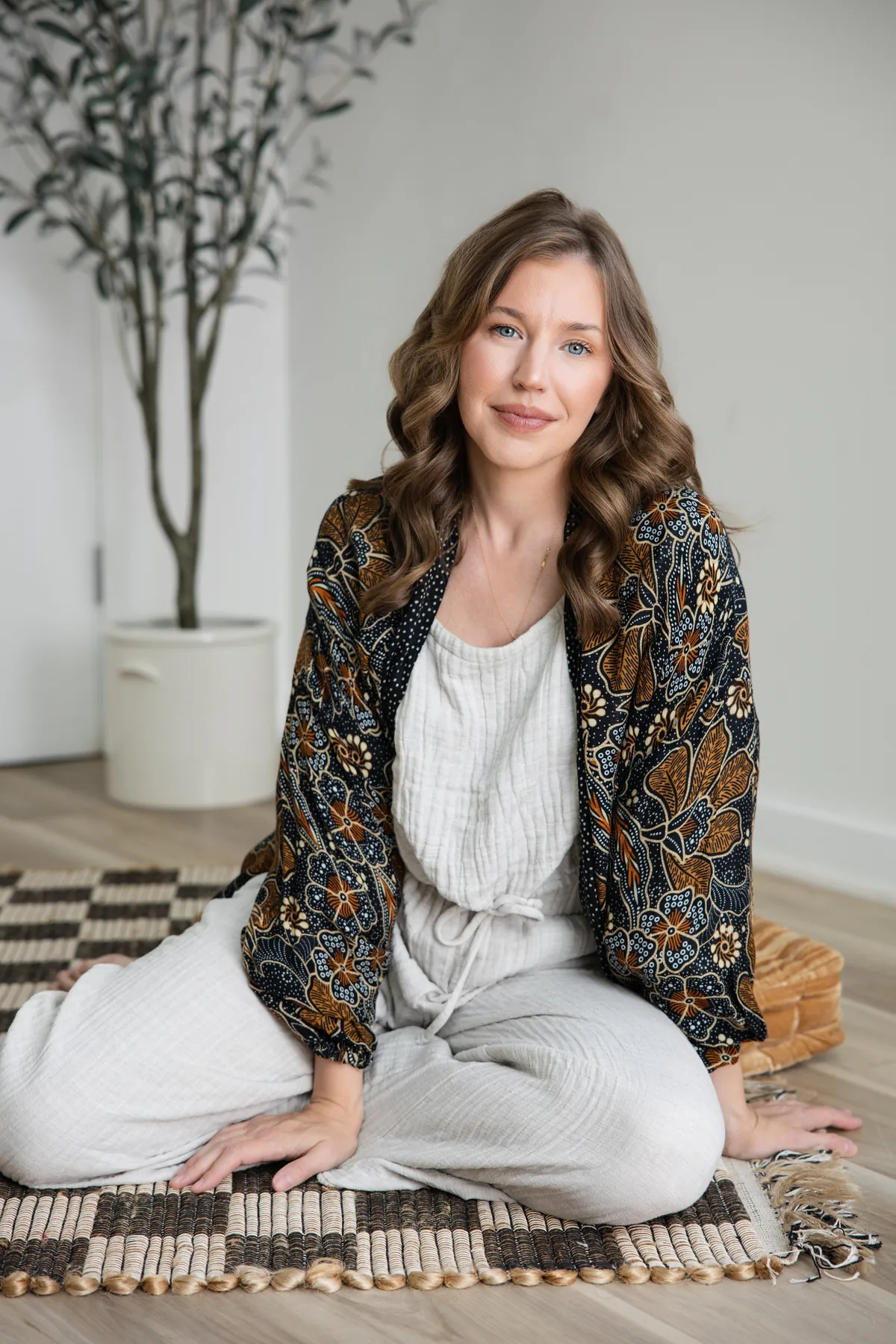 Sarah Bland, founder of Enter Peace Print and Wellness Collective, sitting on a woven rug in a bright room with an olive tree behind her